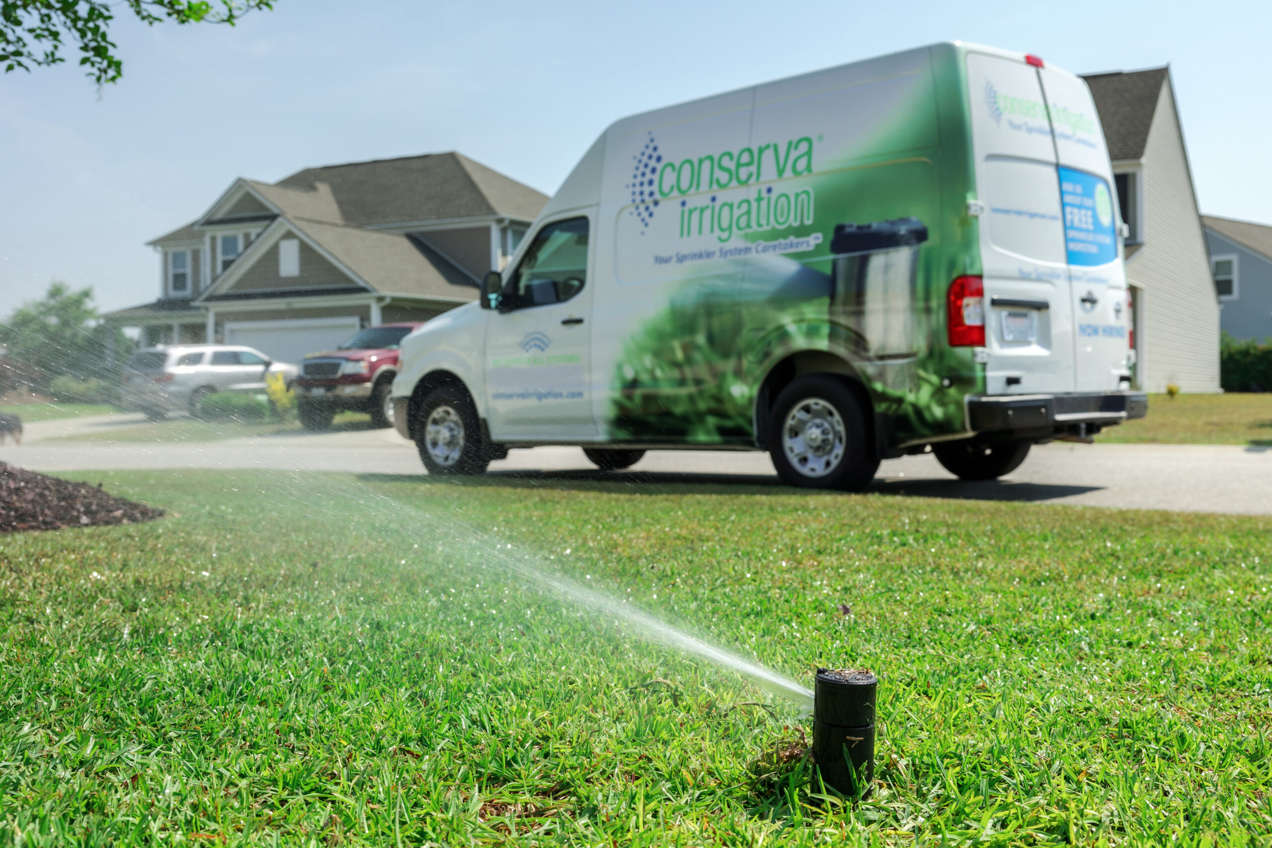 A Conserva Irrigation franchise service van parked on a street, with a sprinkler system actively watering the lush green lawn in the foreground.